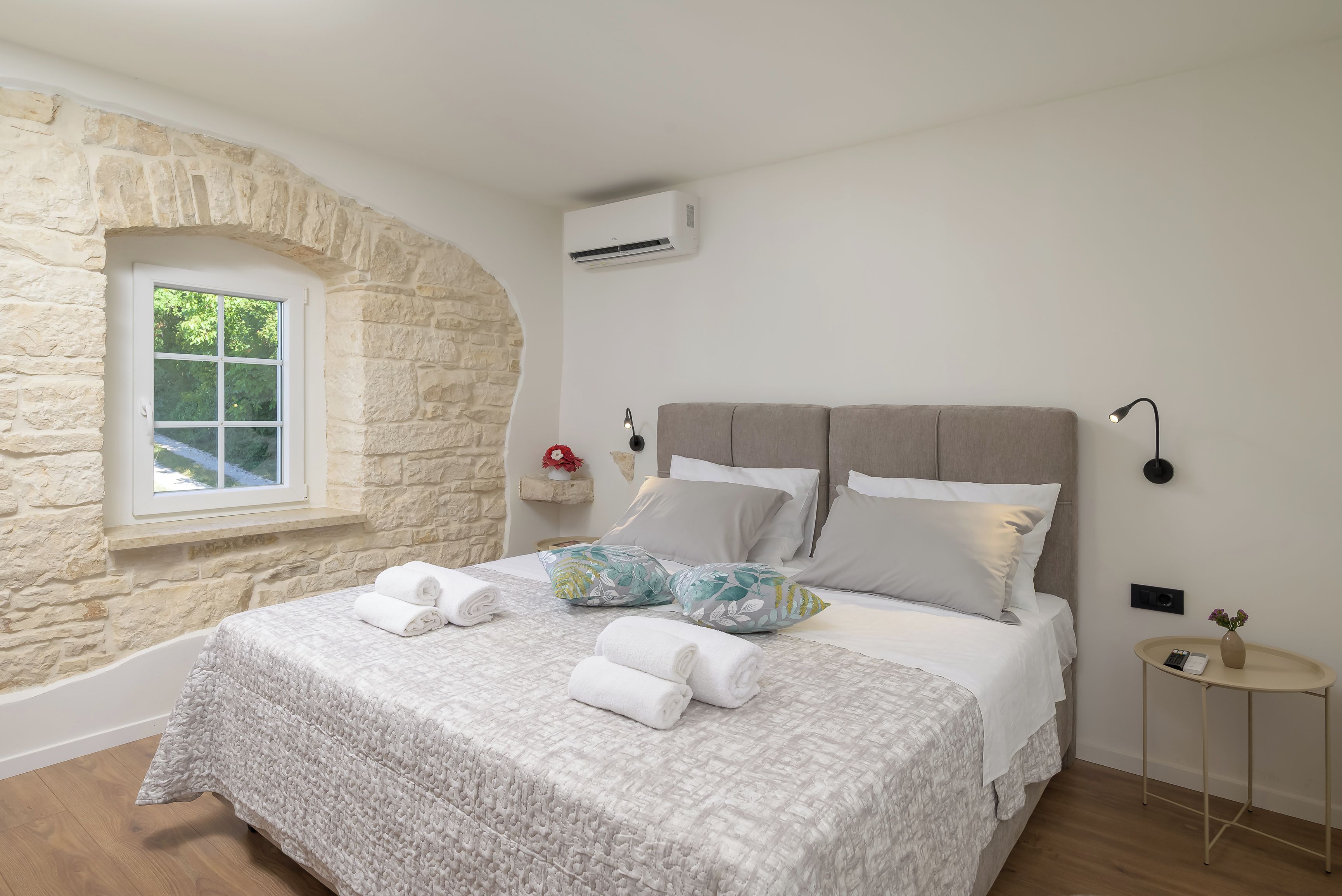 A bedroom with a stone wall, a window, a bed with white bedding, and a bedside table with a vase.