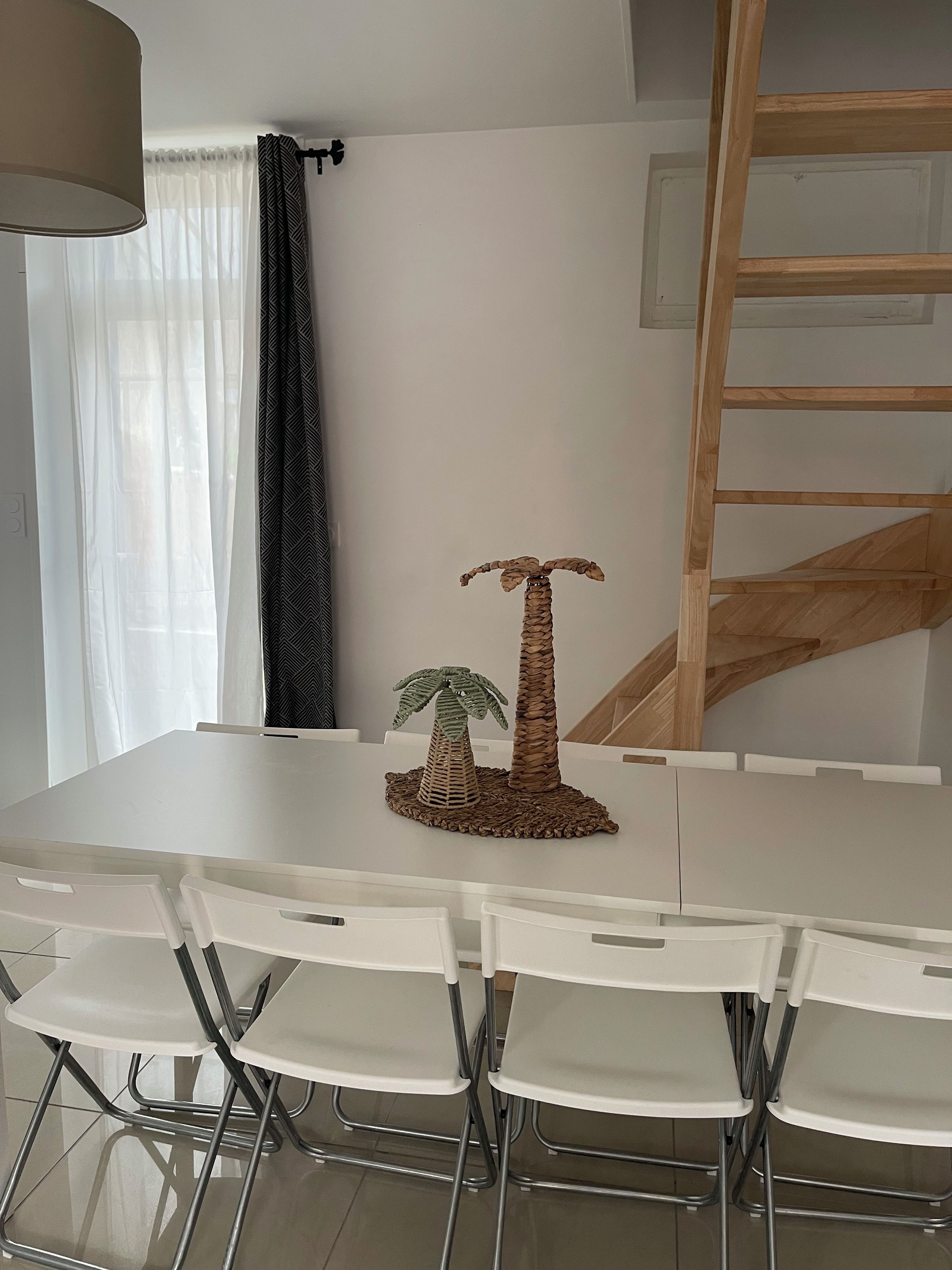 Un coin repas avec une table blanche, des chaises blanches, une décoration centrale faite d’une composition de feuilles de palmier tressées et une échelle en bois appuyée contre le mur.