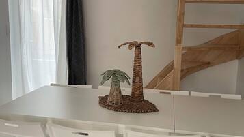 Un coin repas avec une table blanche, des chaises blanches, une décoration centrale faite d’une composition de feuilles de palmier tressées et une échelle en bois appuyée contre le mur.