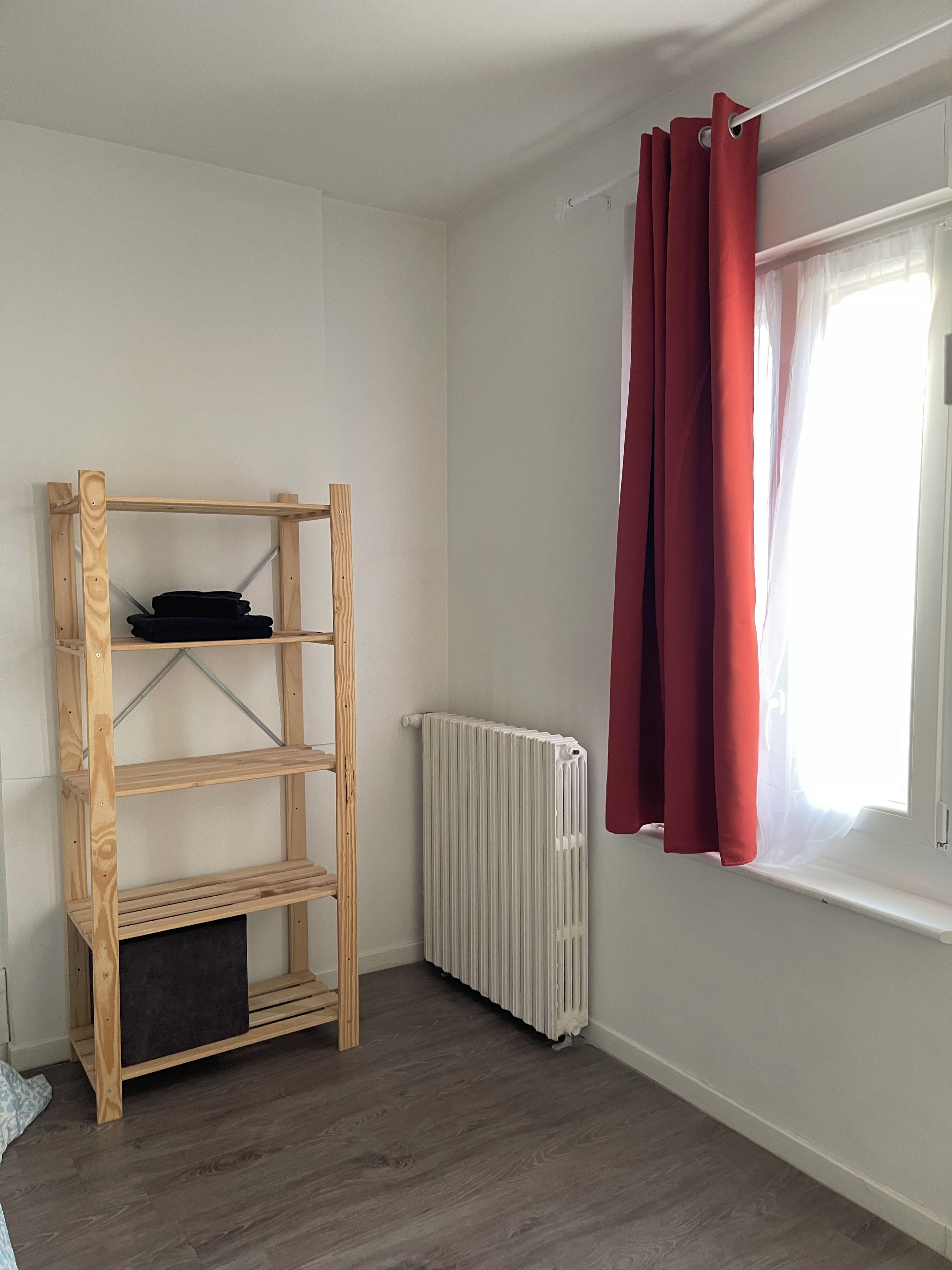 A room with a wooden shelving unit, a radiator, and a window with red curtains.