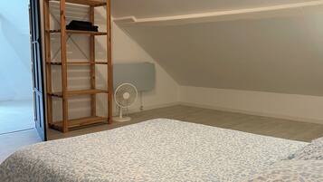 Une chambre avec un plafond en pente, un lit recouvert d’une couvre-lit à motifs, une étagère en bois et un ventilateur sur pied.