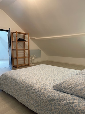 A bedroom with a sloped ceiling, a bed with a patterned bedspread, a wooden shelf unit, and a standing fan.