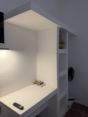 Un estante blanco con un control remoto y libros, un ventilador de pared y un estante con cestas.