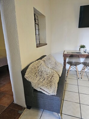 Una habitación con un sofá gris, una ventana pequeña, una mesa de madera con una planta y un suelo de baldosas.