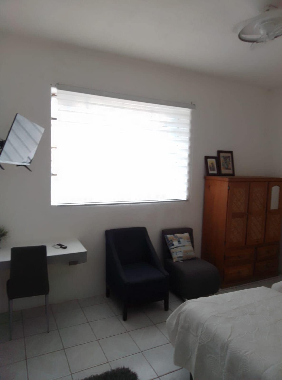 Une petite pièce bien éclairée, avec un bureau, une chaise, un lit et une fenêtre équipée de stores.