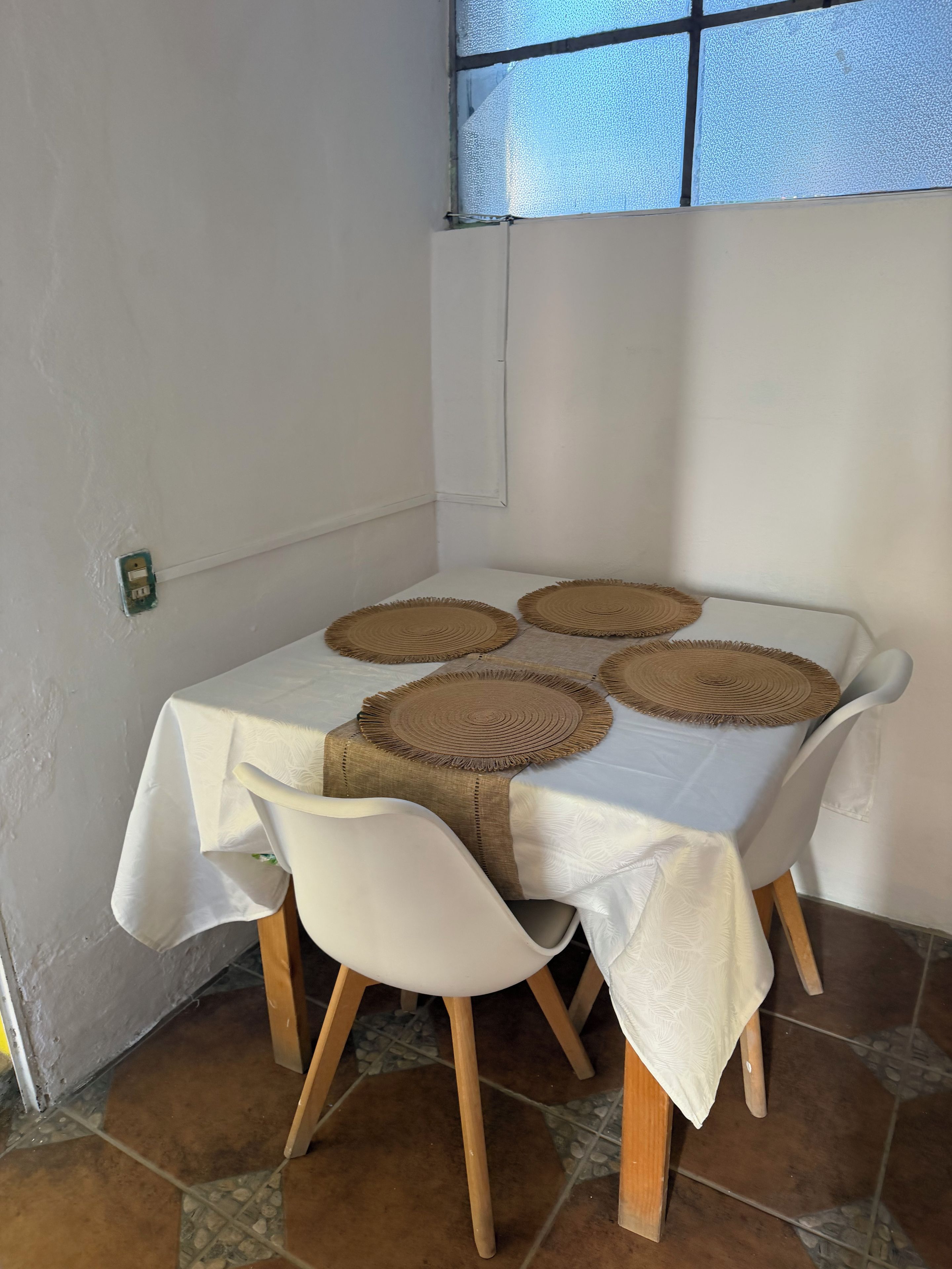 Una mesa con mantel blanco, cuatro individuales tejidos y dos sillas blancas con patas de madera.