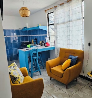 Un espacio habitable compacto con kitchenette, una zona de comedor con sillas azules y un sofá con cojines amarillos.