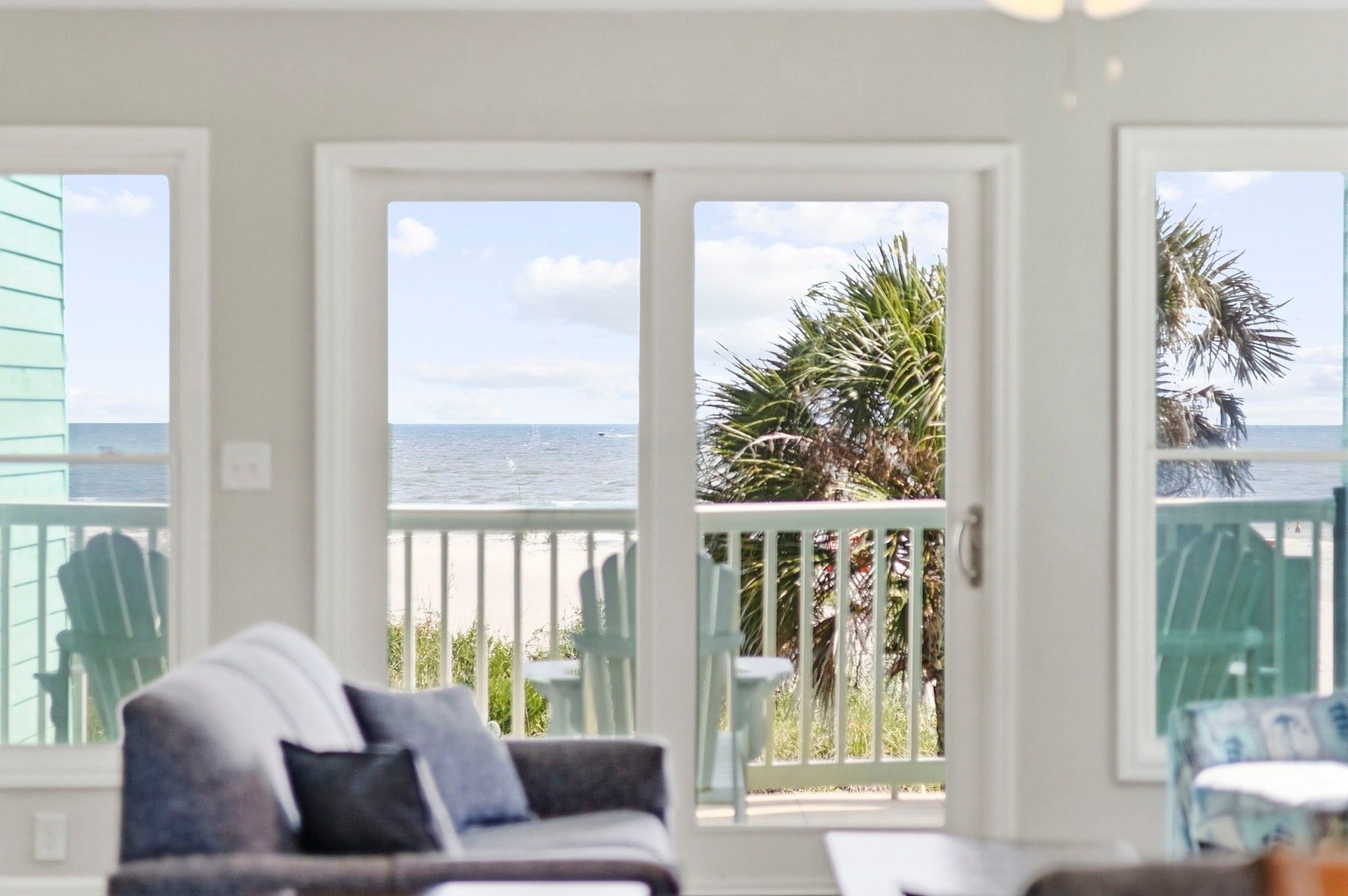 A room with a view of the ocean, a balcony with chairs, and a door leading outside.