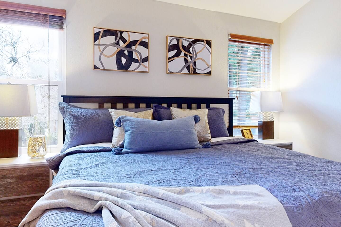 A bedroom with a large bed, two wall-mounted abstract paintings, and a window with blinds.