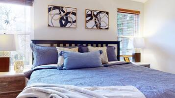 A bedroom with a large bed, two wall-mounted abstract paintings, and a window with blinds.