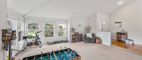 Ein geräumiger Raum mit Kicker, Fitnessgeräten und Blick auf Grünanlagen durch große Fenster.
