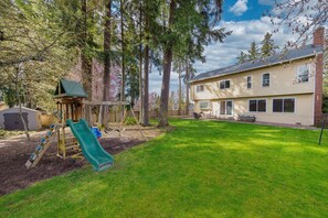 A backyard with a wooden playset and a green slide, a grassy lawn, and a two-story building with a brick chimney. - Tech-Ready Suite @ Education Hill | Fast WiFi & AC (Redmond)