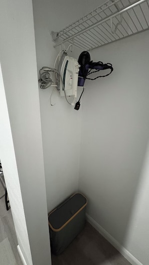 Eine Ecke eines Raumes mit weißer Wand, einer an der Decke befestigten Ablage, einem Staubsauger und einem Mülleimer.