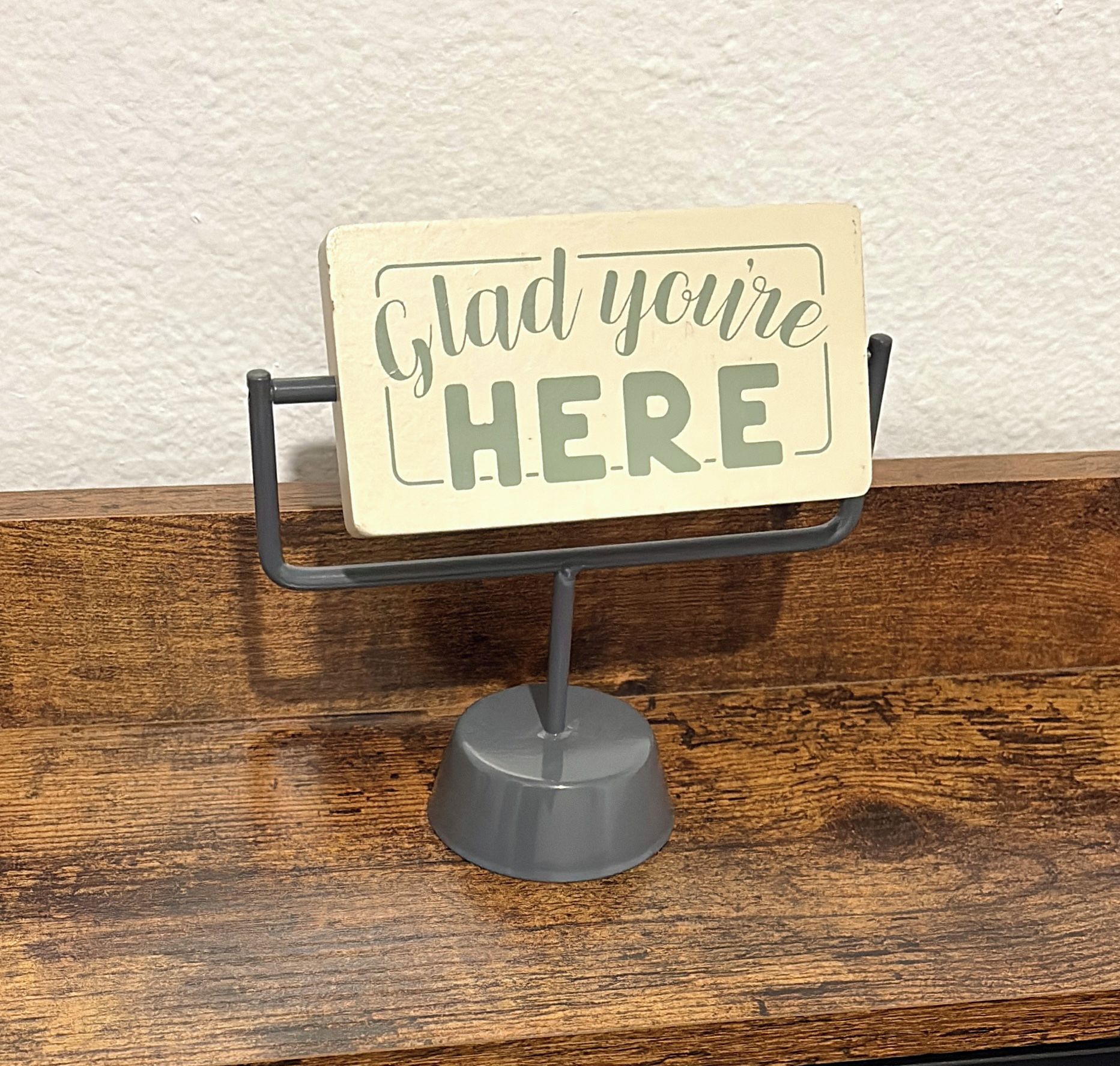 Un letrero sobre un soporte con el texto 'Glad you're HERE' frente a un fondo de pared texturizada.