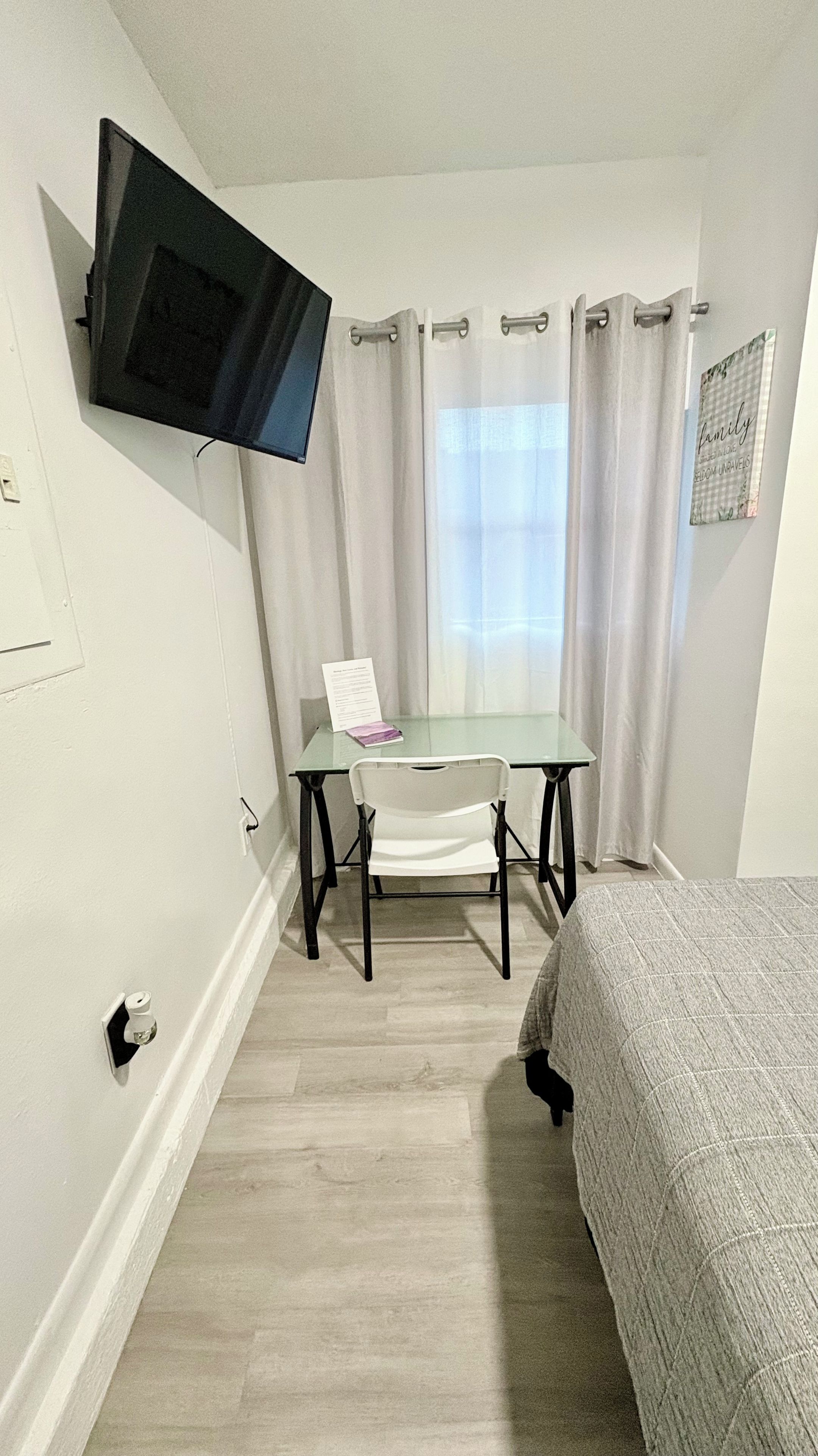 A room with a bed, a table with a chair, a TV mounted on the wall, and a window with curtains.
