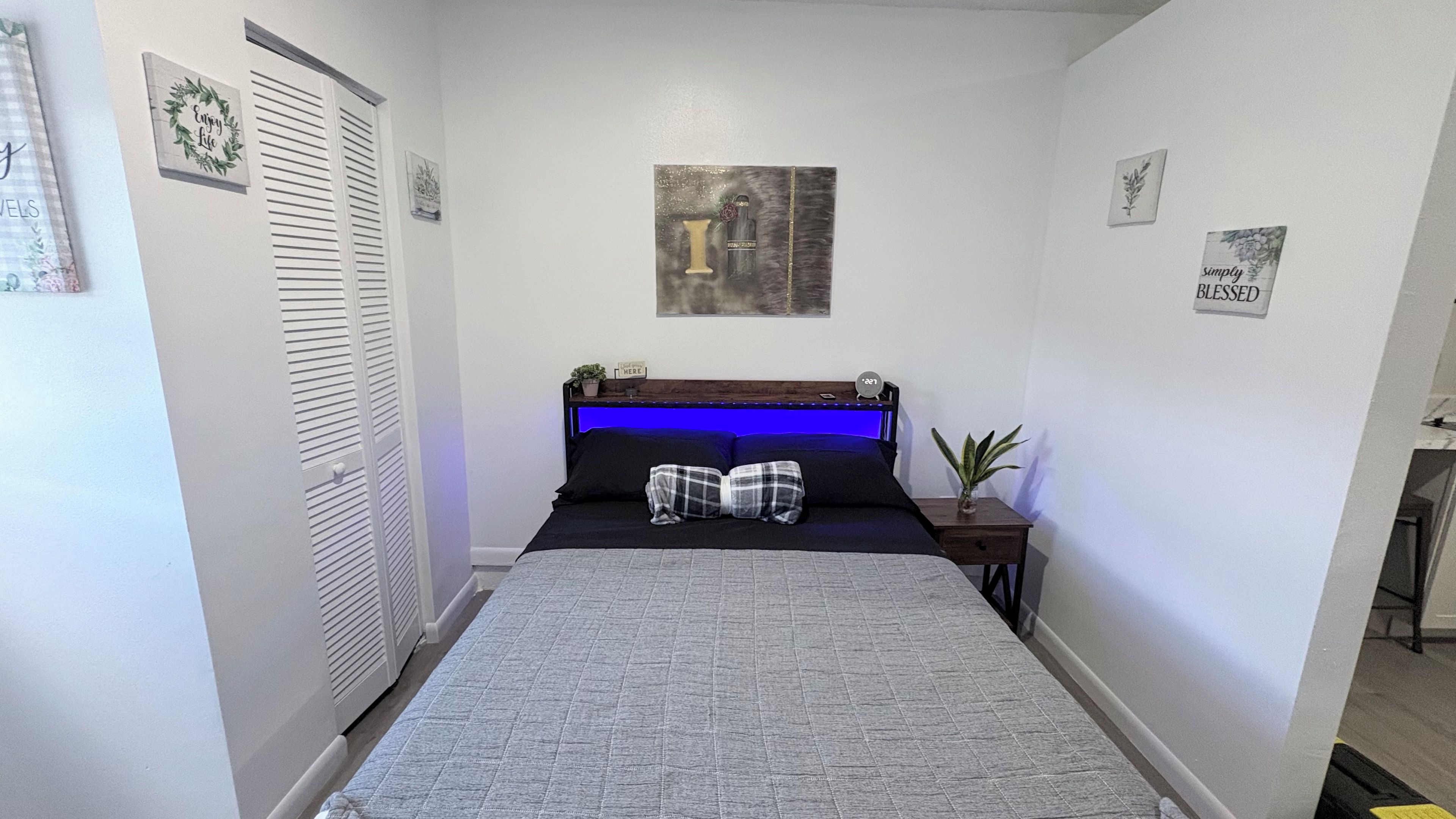 A bedroom with a bed, a bedside table, a wall clock, and a small potted plant.