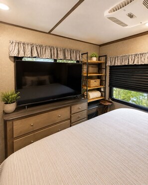 A bedroom with a bed, a TV, a dresser, a shelf with baskets, and a window with curtains. - Rivers Edge Retreat at River Rock Landing (Gadsden)
