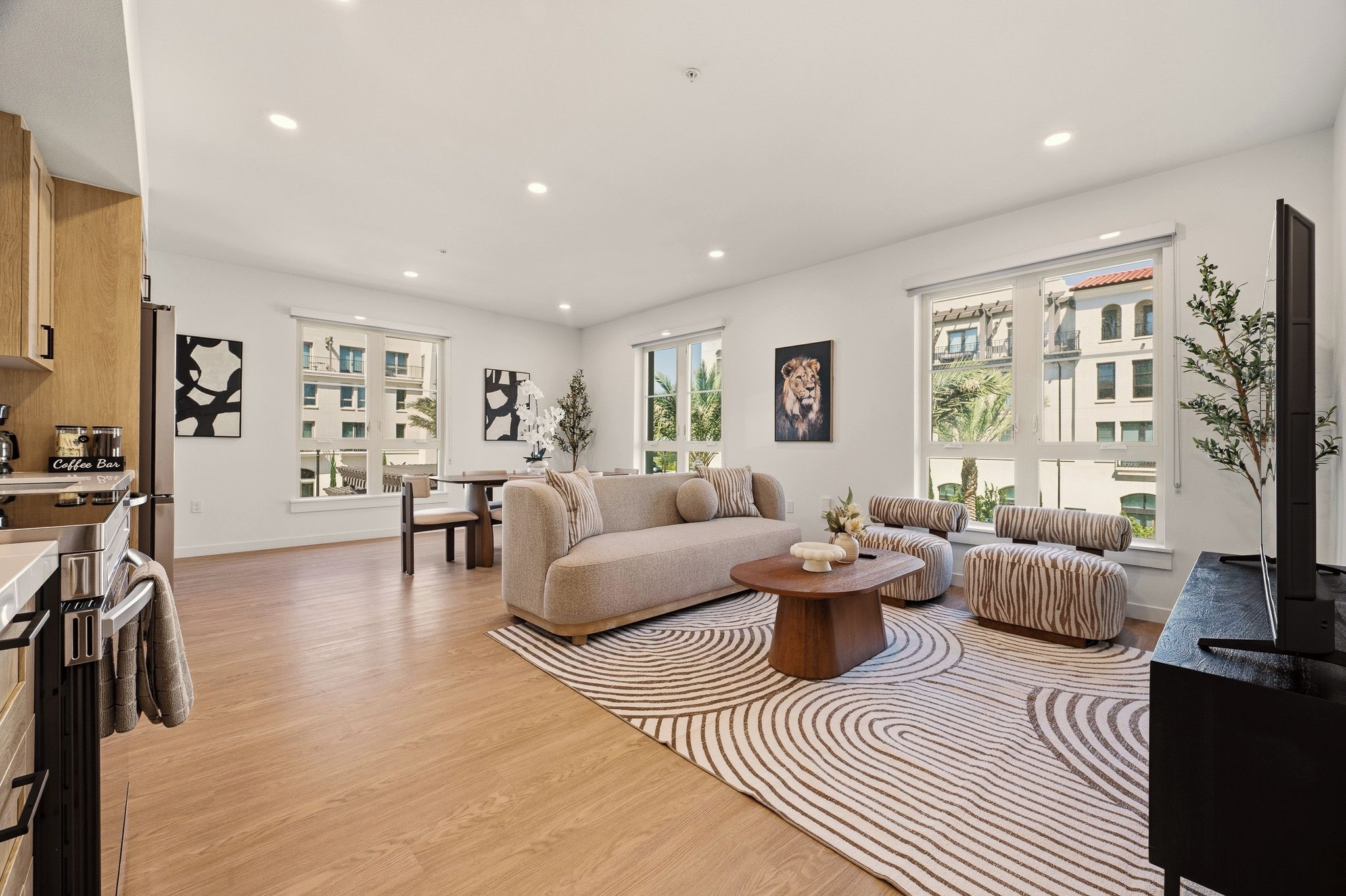 A spacious living area with a sofa, ottoman, coffee table, and a TV. There are windows providing natural light and a view of buildings outside.