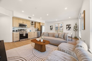 A modern living room with a sofa, coffee table, and kitchen area with appliances. - Modern King 2B2B｜Pool & Gym｜70in Smart 4K TV (Montclair)