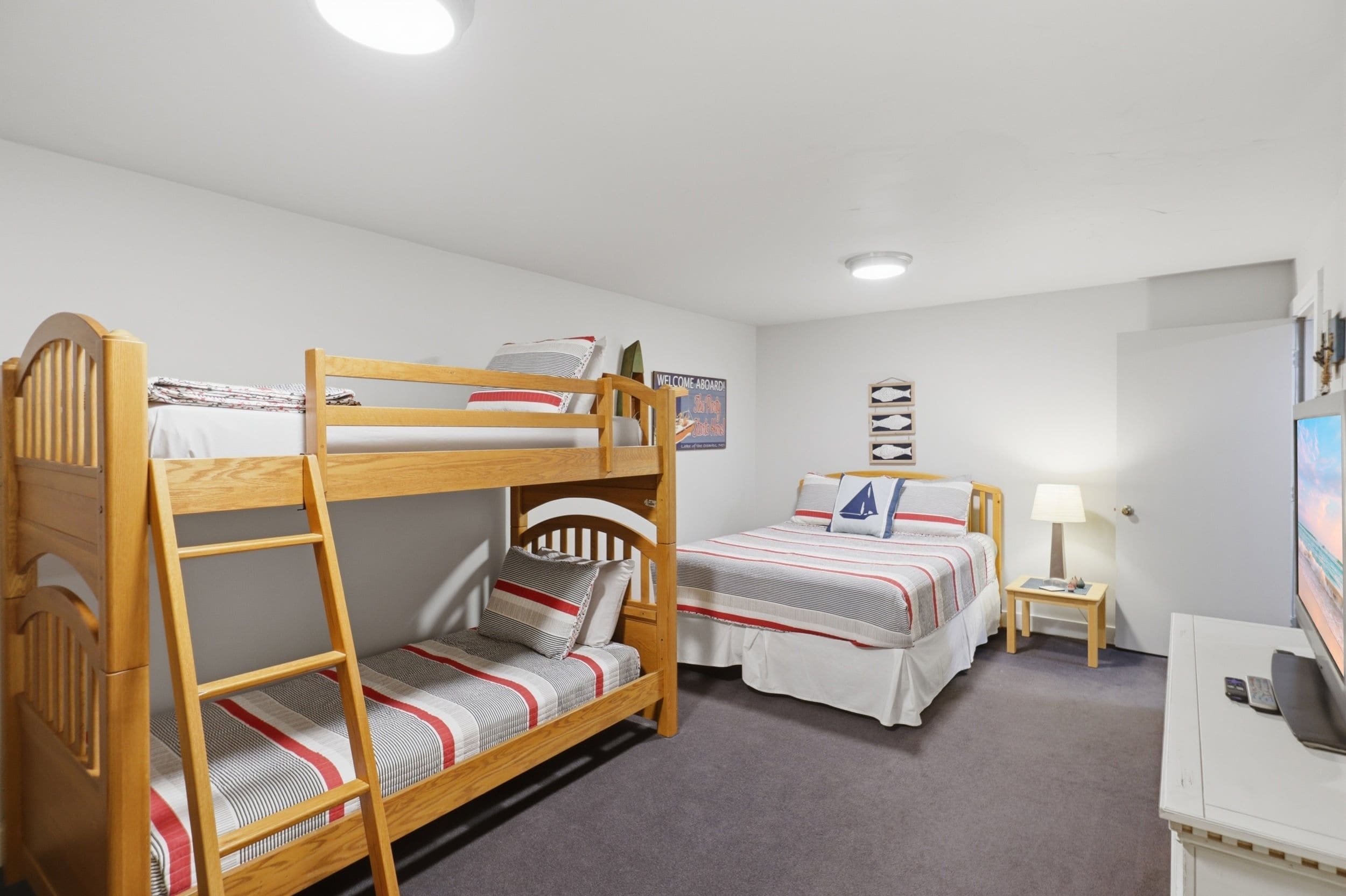 A bunk bed room with a desk and a chair, a small table with a lamp, and a wall-mounted TV.