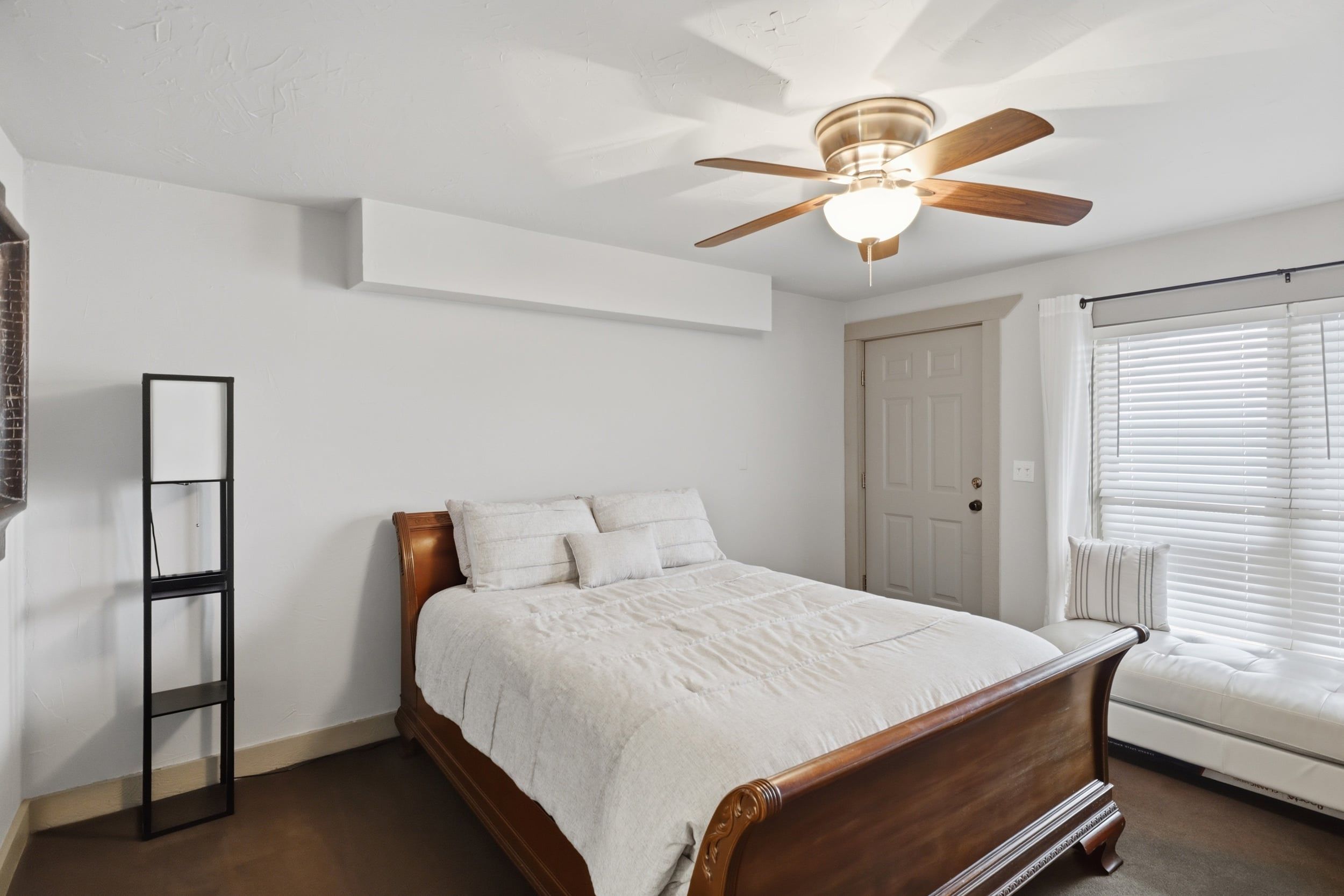 Een slaapkamer met een houten bed, een plafondventilator, een witte muur en een raam met zonwering.