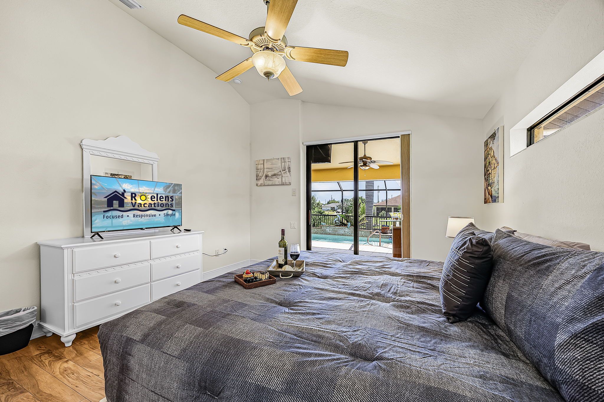 A bedroom with a bed, a TV on a dresser, a ceiling fan, and a view of the outdoors.