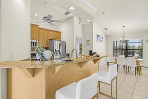 Cozinha moderna com ilha central, armários de madeira, eletrodomésticos de aço inoxidável e área de jantar com cadeiras brancas e lustre.