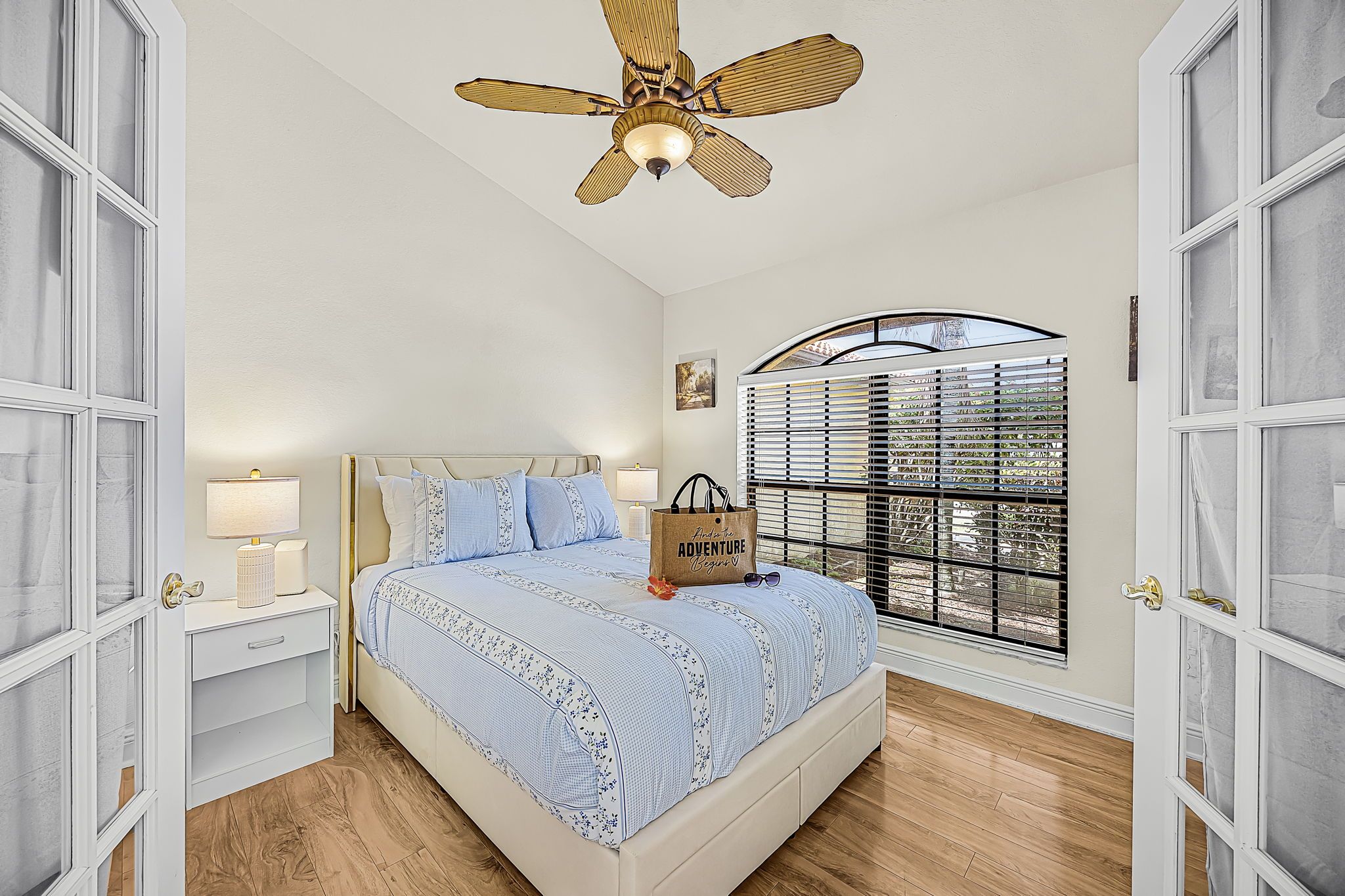 Une chambre à coucher comprenant un lit, une table de chevet, une lampe, une fenêtre avec des stores et un ventilateur de plafond.