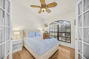 Quarto com cama, mesa de cabeceira, luminária, janela com persianas e ventilador de teto.