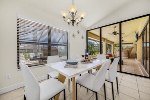 A dining area with a table set for four, white chairs, a chandelier, and a view through a glass door to an outdoor space. - Canal Retreat with Heated Pool, Bikes & Waterfront Fun in Cape Coral - Villa Amarillo - Roelens (Cape Coral)