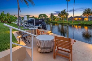 An outdoor seating area with a table, chairs, and a grill, overlooking a calm body of water. - Tropical Canal Escape with Pool, Spa & Kayaks in Cape Coral - Pineapple Princess - Roelens (Cape Coral)