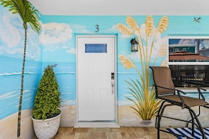 A door with a number 3, a potted plant, a chair, and a wall mural with a beach scene. - Poolside Fun, Pickleball & Coastal Adventures Await in Fort Myers - Jefferson Palms - Roelens (Fort Myers Beach)