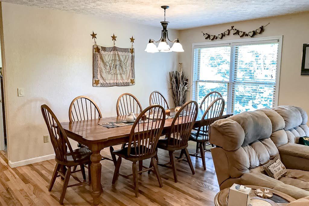 A dining area with a wooden table and chairs, a chandelier, and a window with blinds.