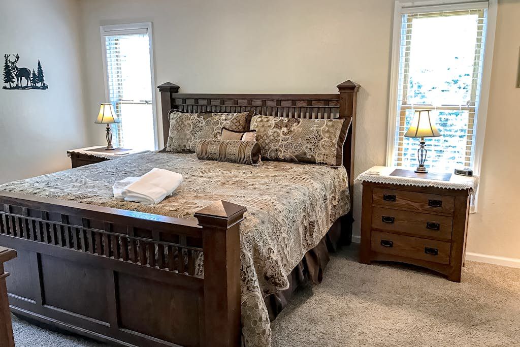 Un dormitorio con cama de madera, dos mesitas de noche con lámparas, un adorno en la pared y una ventana con persianas.