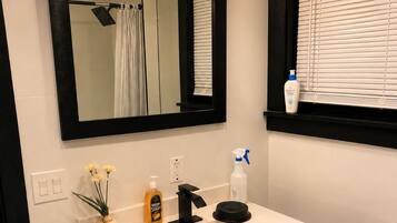 A bathroom with a white sink, black vanity, a mirror with lights, a window with blinds, and a teal bath mat.