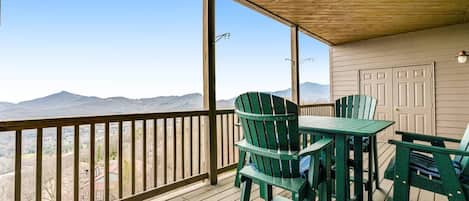 A covered outdoor area with a wooden deck, green Adirondack chairs, and a view of distant mountains.
