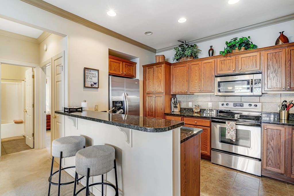 A kitchen with wooden cabinets, a stainless steel refrigerator, an oven, a microwave, and a bar area with stools.