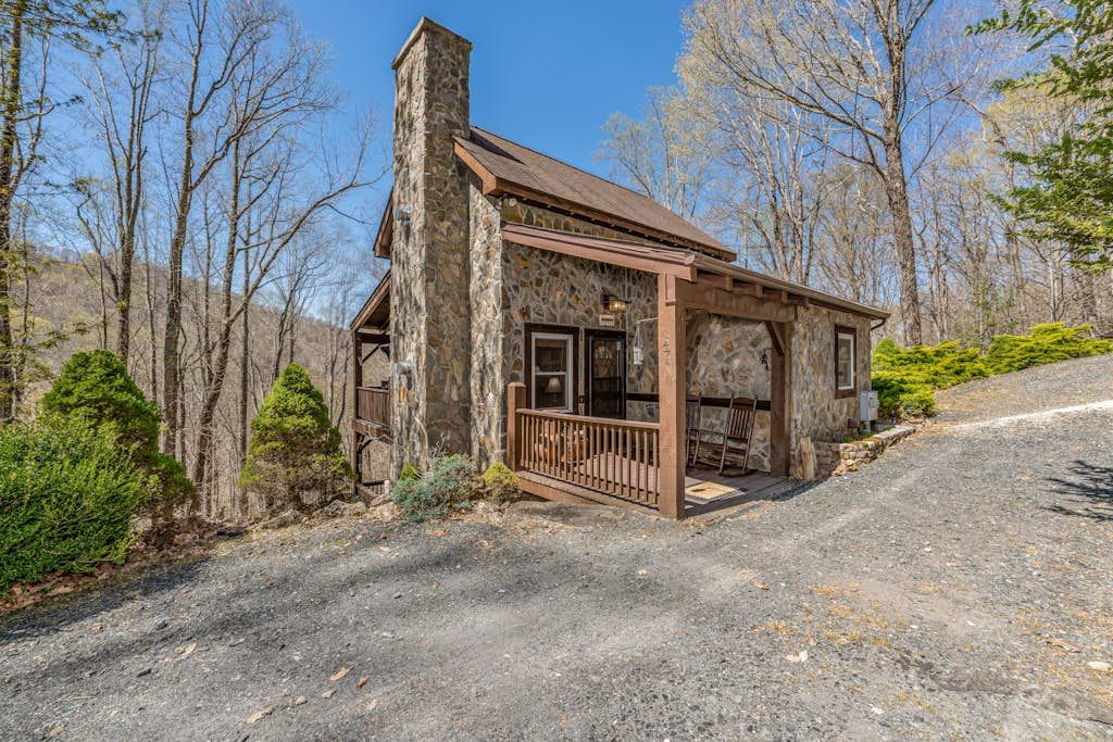 A rustic stone building with a wooden porch, surrounded by trees and a gravel path.