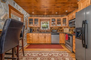 A kitchen with wooden cabinets, a refrigerator, a dishwasher, and a stone wall. - Mountainside Charm: Cozy cabin near Banner Elk with 3 fireplaces, deck swing bed (Elk Park)