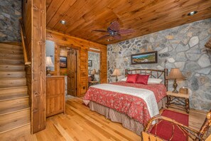 A wooden cabin bedroom with a stone wall, a bed with floral bedding, a wooden chair, and a ceiling fan. - Mountainside Charm: Cozy cabin near Banner Elk with 3 fireplaces, deck swing bed (Elk Park)