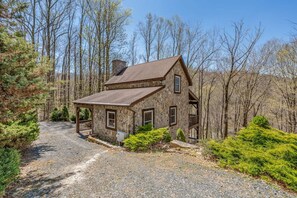 A stone house with a sloped roof, a wooden porch, and a gravel driveway surrounded by trees. - Mountainside Charm: Cozy cabin near Banner Elk with 3 fireplaces, deck swing bed (Elk Park)