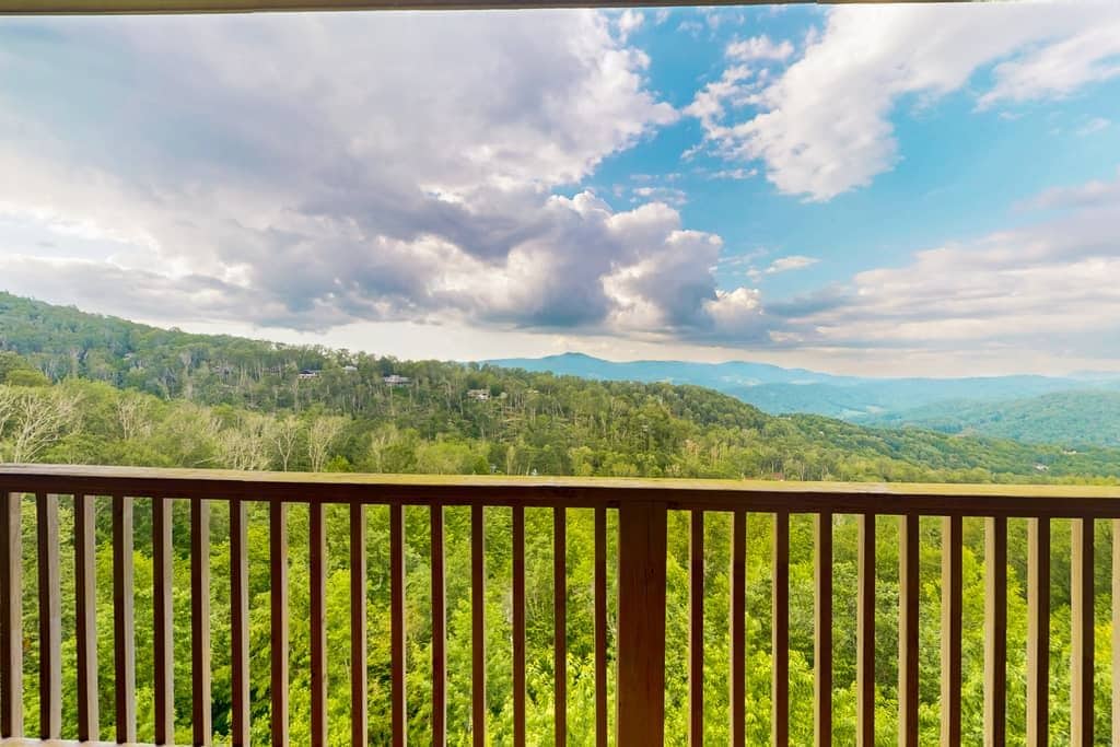 A view from a balcony with a railing, overlooking a forested landscape with distant mountains under a partly cloudy sky.