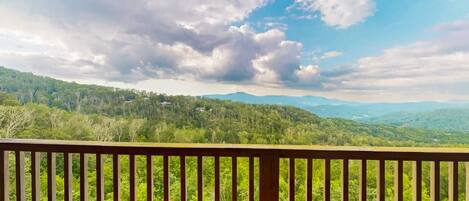 A view from a balcony with a railing, overlooking a forested landscape with distant mountains under a partly cloudy sky.