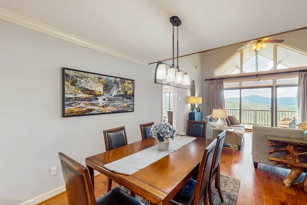 A dining area with a wooden table and chairs, a painting on the wall, and a view of the outdoors through large windows.