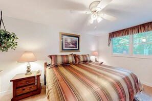 Ein Schlafzimmer mit einem Bett, Nachttischlampen, einem Deckenventilator, einem gerahmten Bild an der Wand und einem Fenster mit Vorhängen.