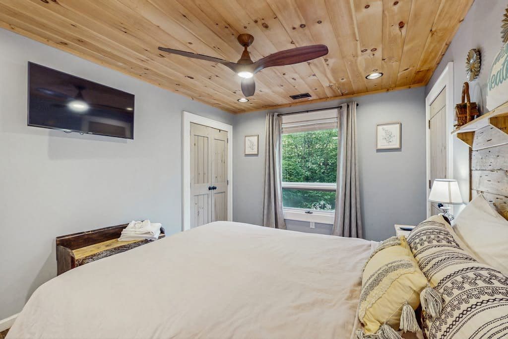 A bedroom with a wooden ceiling, a bed with white bedding, a television on the wall, and a window with curtains.