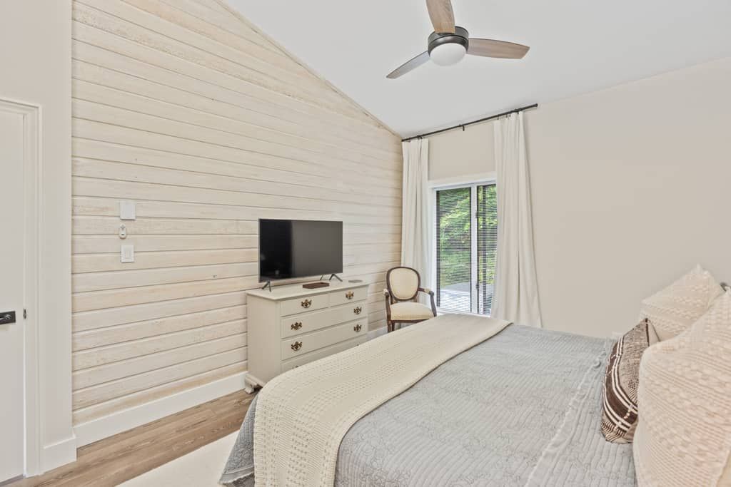 A bedroom with a bed, a television on a dresser, a ceiling fan, and a window with curtains.