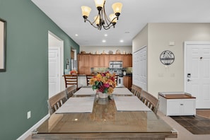 A dining area with a long table, chairs, a chandelier, and a kitchen in the background. - Summit Serenity - Condo on Sugar Mountain with Amazing Views, Gas Fireplace, Walk to Slopes! (Banner Elk)