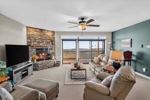 A living room with a stone fireplace, a flat-screen TV, a leather sofa, and a view through a glass door. - Summit Serenity - Condo on Sugar Mountain with Amazing Views, Gas Fireplace, Walk to Slopes! (Banner Elk)