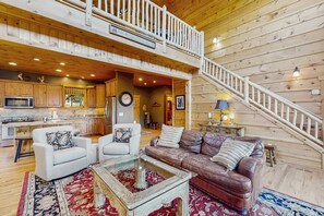 A spacious living area with a high ceiling, wooden flooring, a staircase, and a kitchen in the background. - Howling Rock - Mtn Views, Hot Tub, Pool Table + More (Banner Elk)